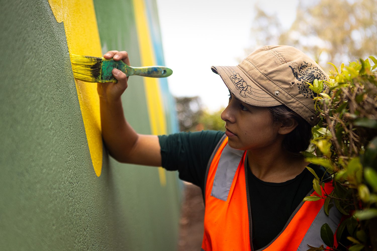 A young woman wearing a cap holds a paint brush as she brushes yellow paint onto a mural