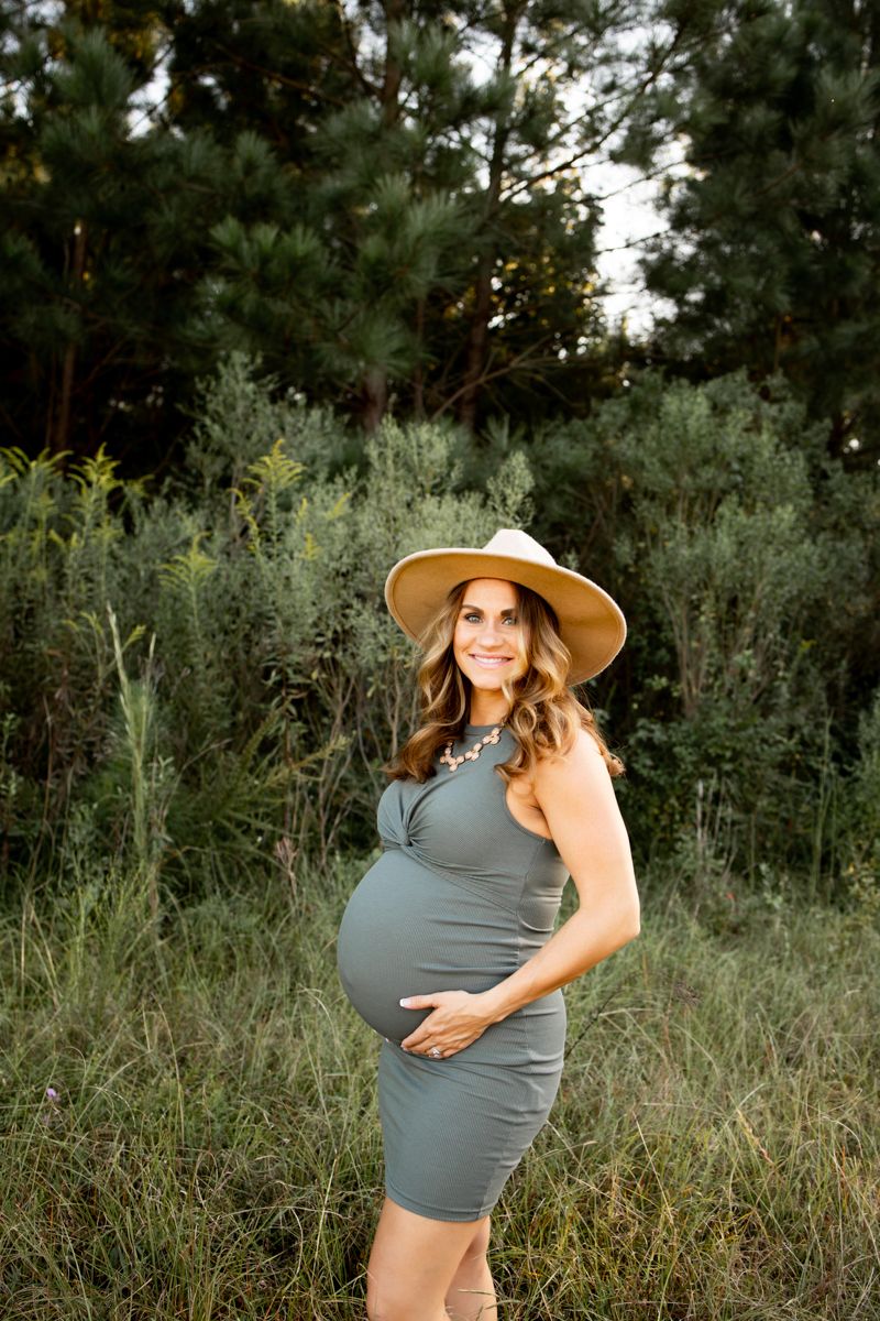 Pregnant lady standing in a grassy field wearing a green dress and brown hat smiling.