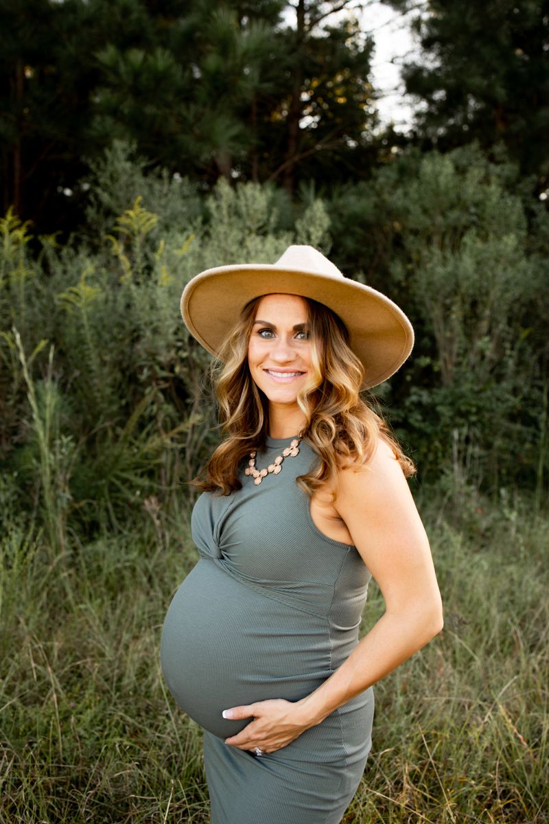Pregnant mother wearing a green dress and brown hat. She is standing in a grassy field cradling her belly smiling.