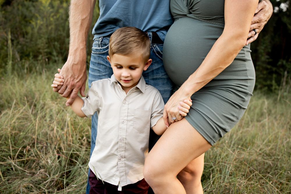 Family of 3 standing in a field focused on little brother.  He is holding mom and dad's hands.