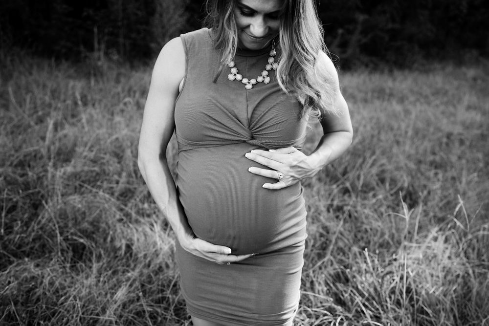Black and white image of pregnant woman standing in a field focusing on her belly while she cradles her belly.