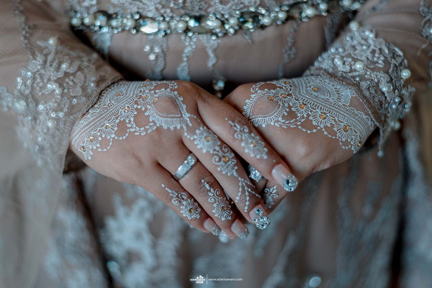 Close-up of hands with delicate white henna patterns and jewelry against a silver beaded dress.