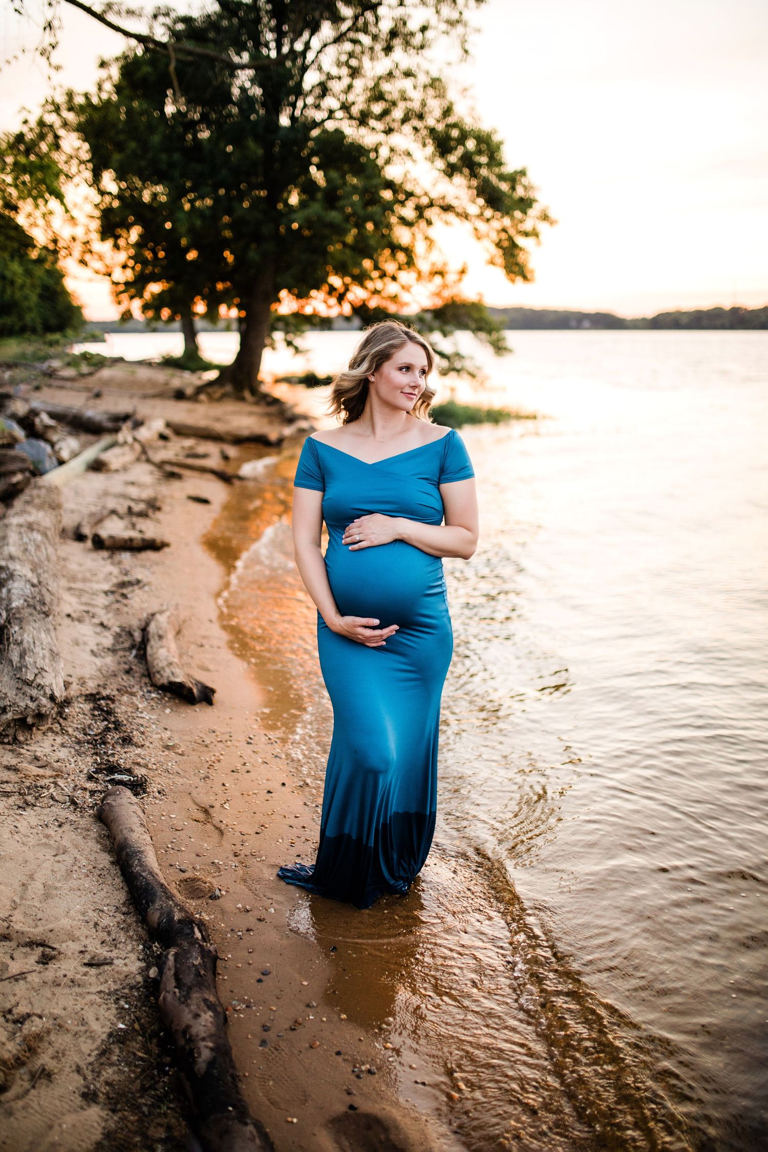 Pregnant woman in blue off-shoulder gown standing in shallow lake water at golden sunset.