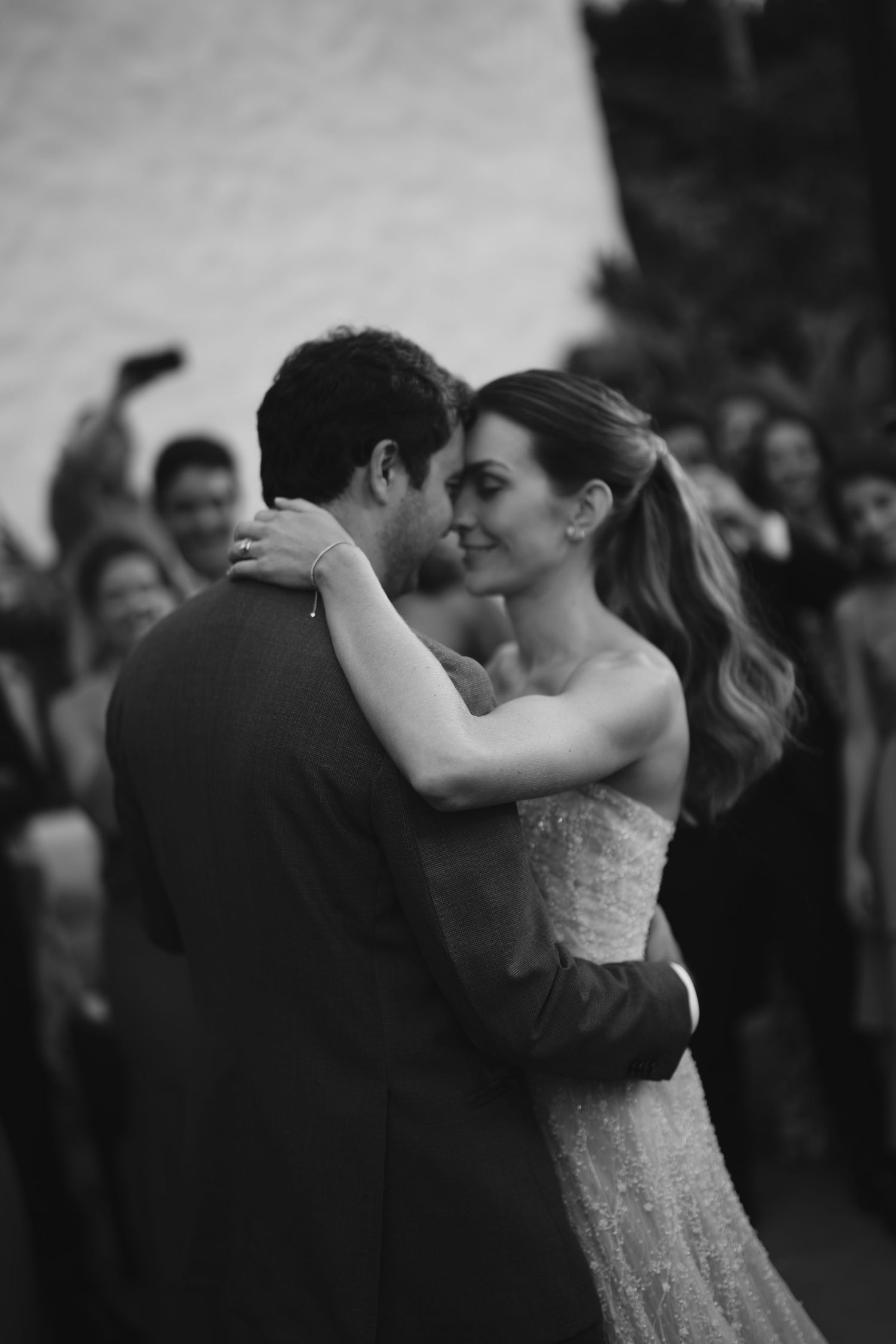 A couple shares their first dance at a wedding reception with guests watching in black and white.