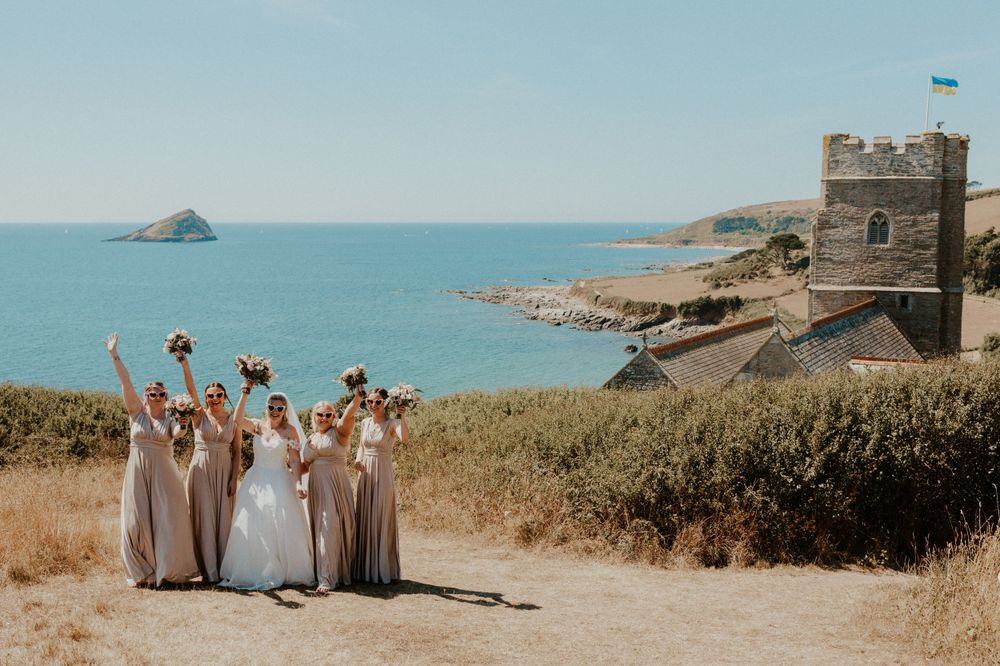 Bride and bridesmaids raise the flowers with the view of Wembury church and the sea behind them