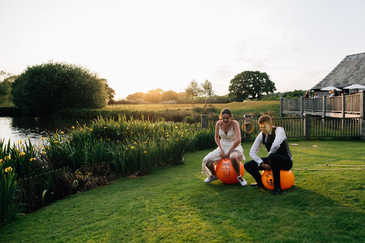Couple sits on orange space hoppers at sunset by scenic pond with garden building.