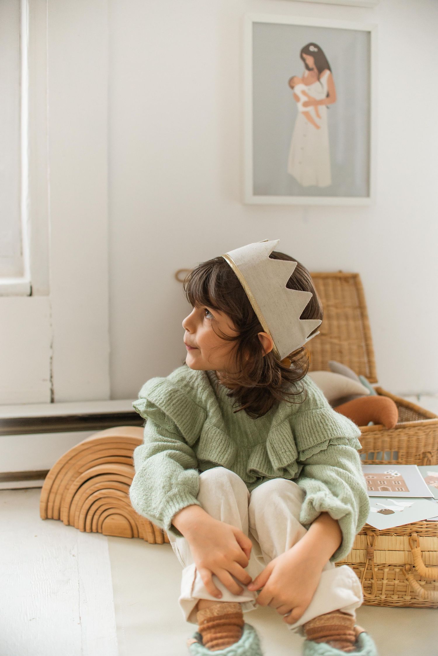 Child wearing a felt crown and mint green sweater sitting on wooden rainbow blocks.