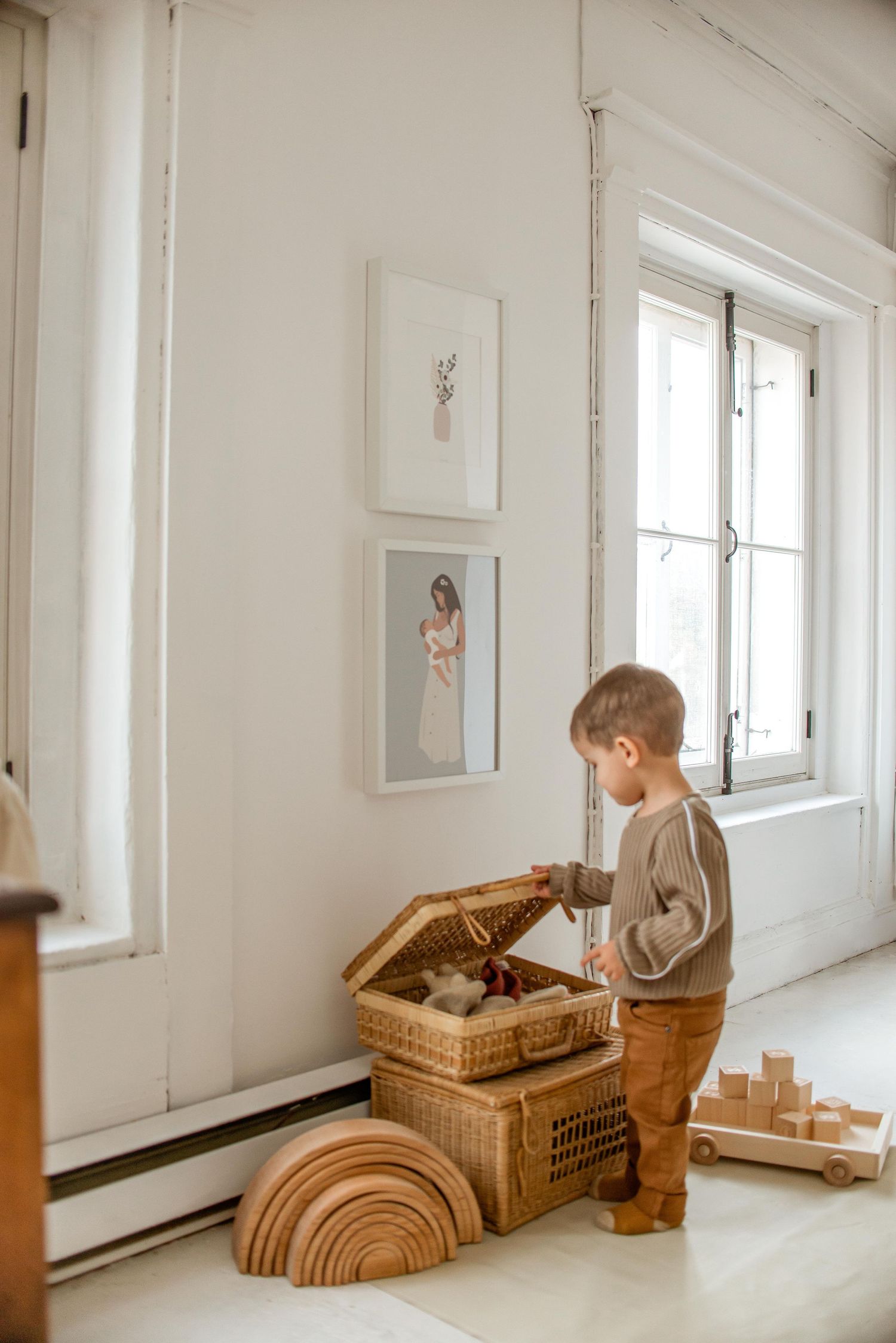 A playroom corner with wooden toys and a woven basket near a bright window with artwork on white walls.