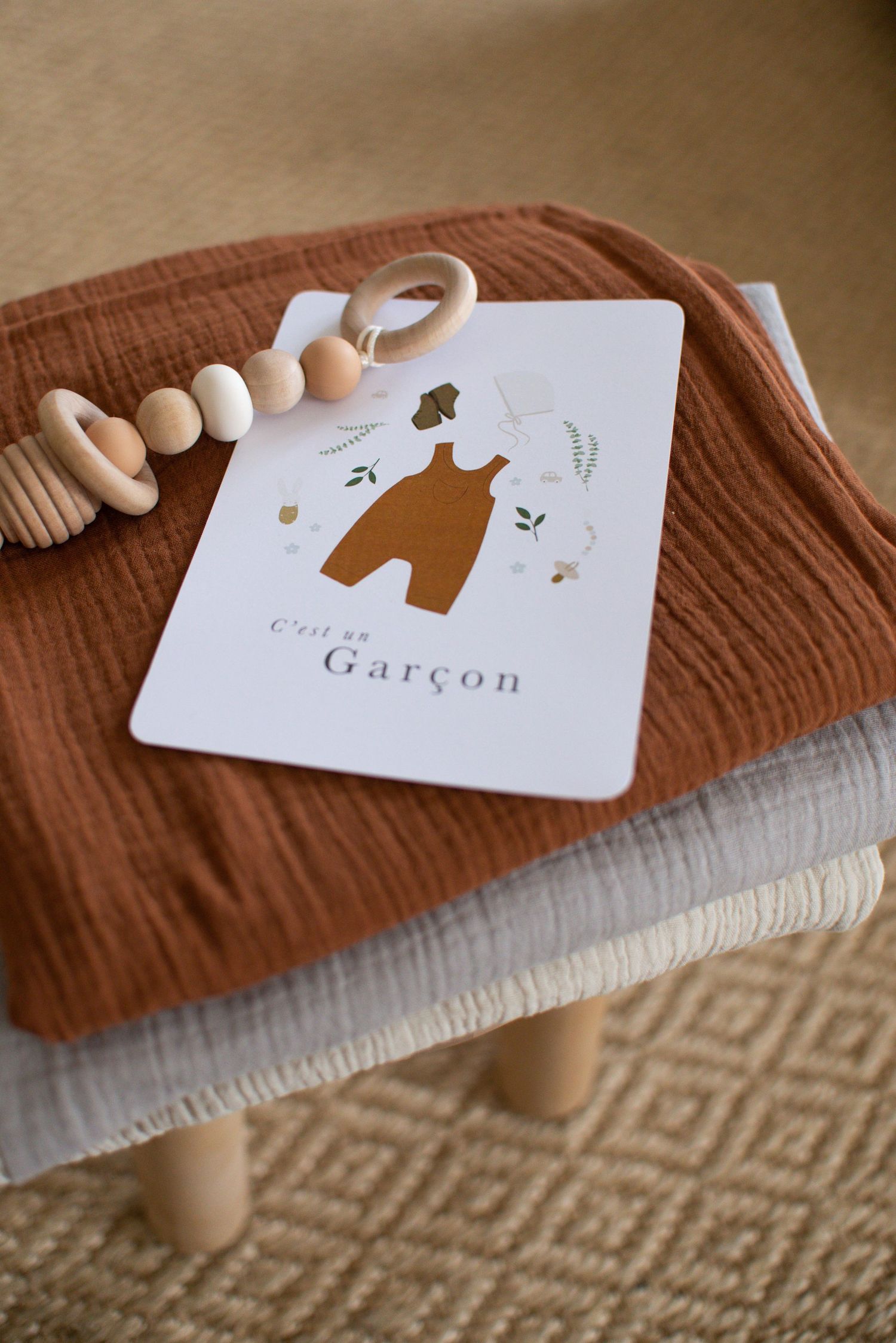 Card design featuring brown bear illustration and 'Garcon' text on textured fabric with wooden beads.