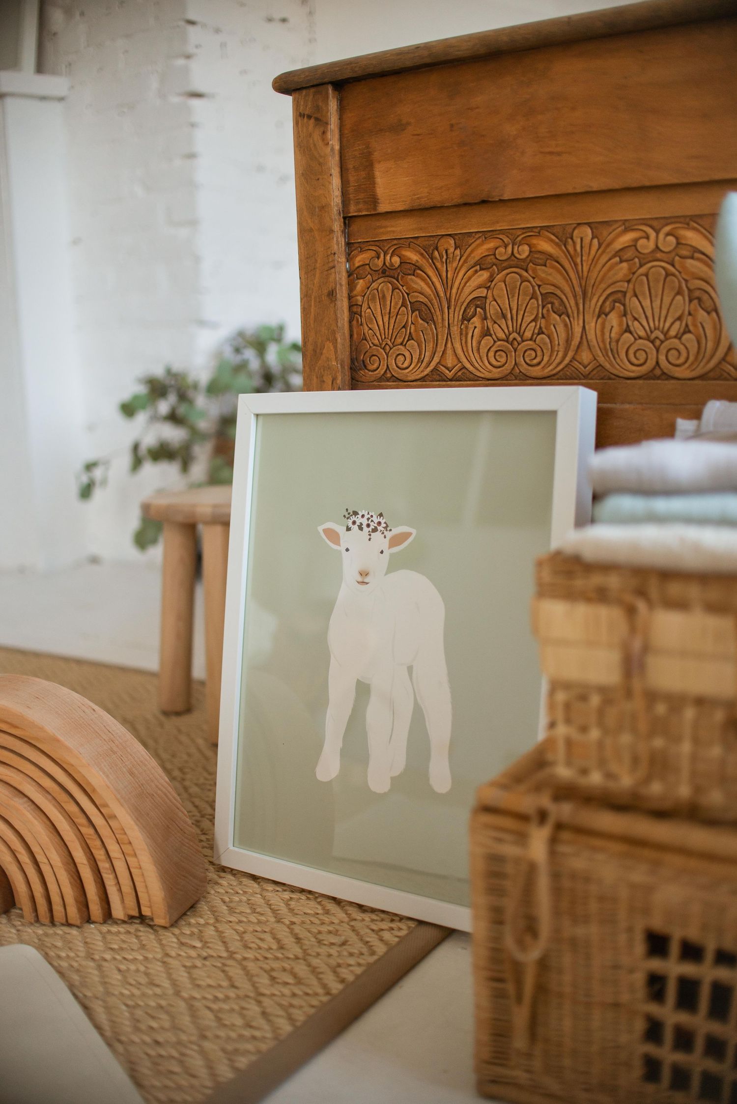 Framed nursery art print of a white lamb with floral crown displayed against carved wooden furniture and decor.
