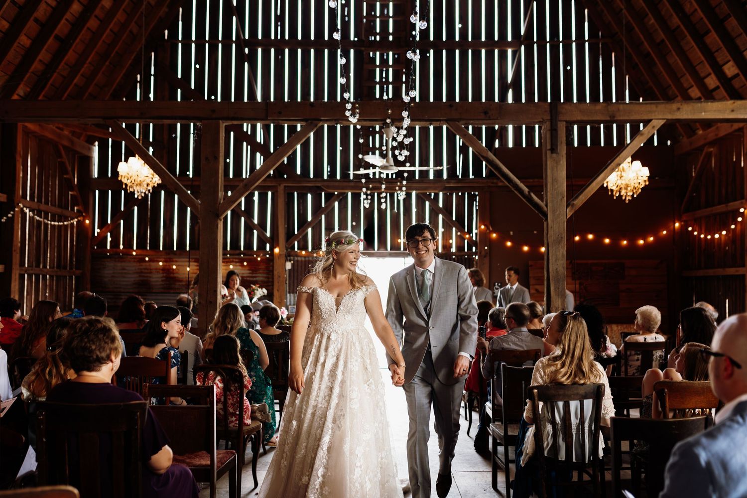 Couple walks down the aisle of a rustic barn wedding venue decorated with string lights and wooden beams.