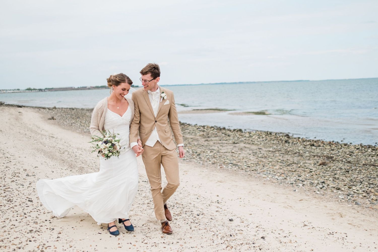 A couple walks along a scenic beach shoreline with flowing white dress and tan suit against the ocean backdrop.