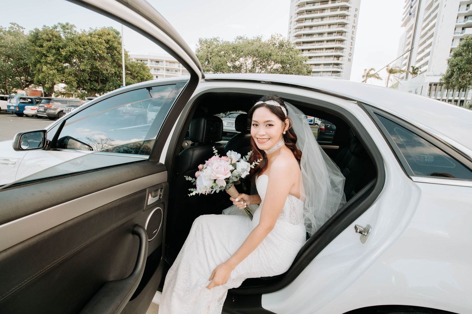 A bride in a white wedding dress sits in a car holding a bouquet.