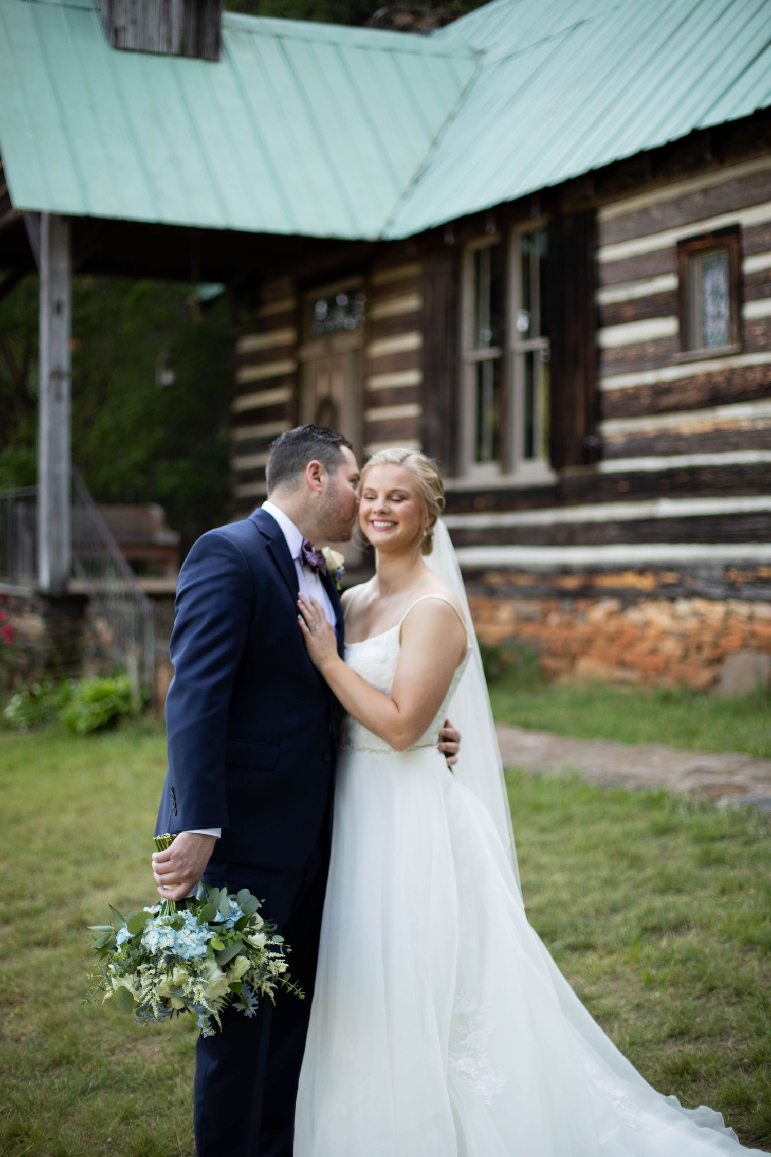 Newlyweds share intimate moment outside rustic log cabin with bride holding blue bouquet.