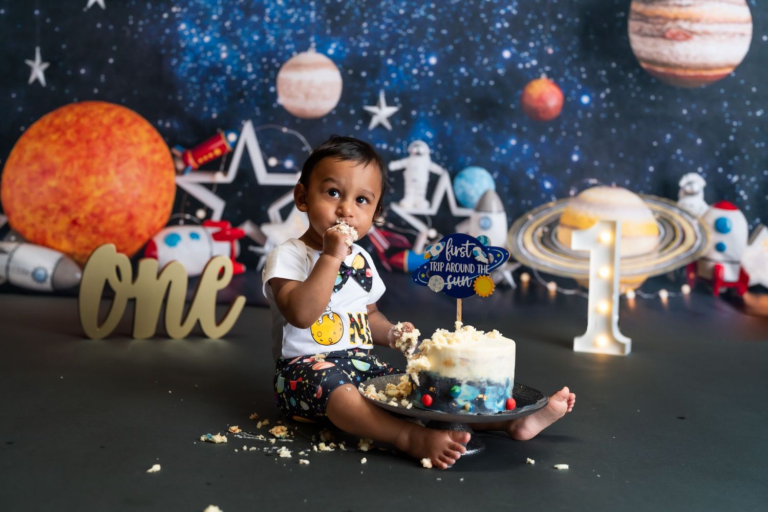Baby in space-themed cake smash photo session with planets and stars backdrop.