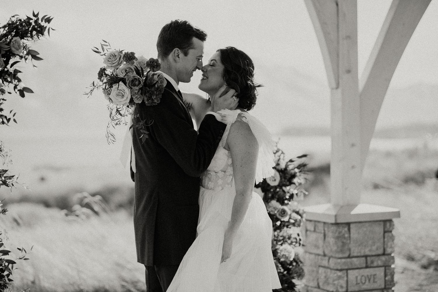 Black and white photo of wedding couple embracing under outdoor pergola with floral bouquet.