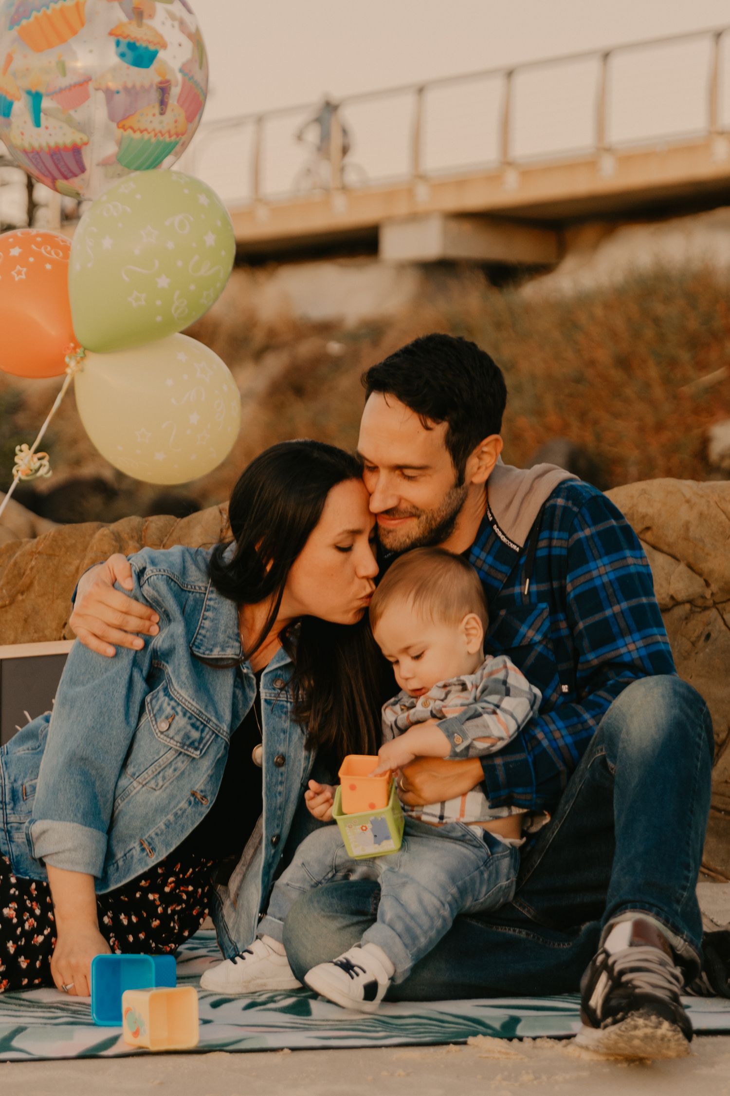 Family picnic outdoors with colorful balloons as parents cuddle with baby on blanket.