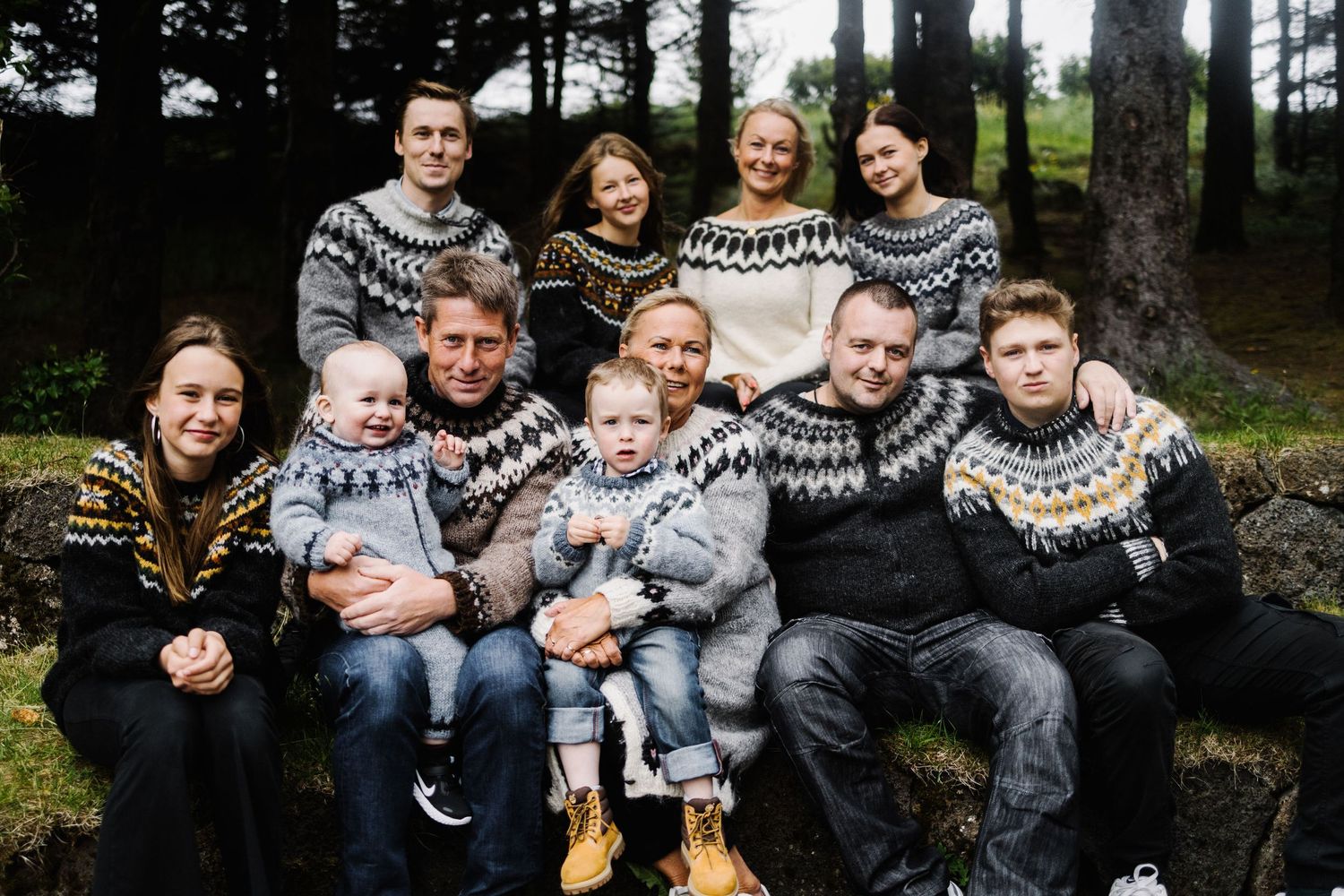 Large family group wearing matching Nordic sweaters posed together outdoors among pine trees.