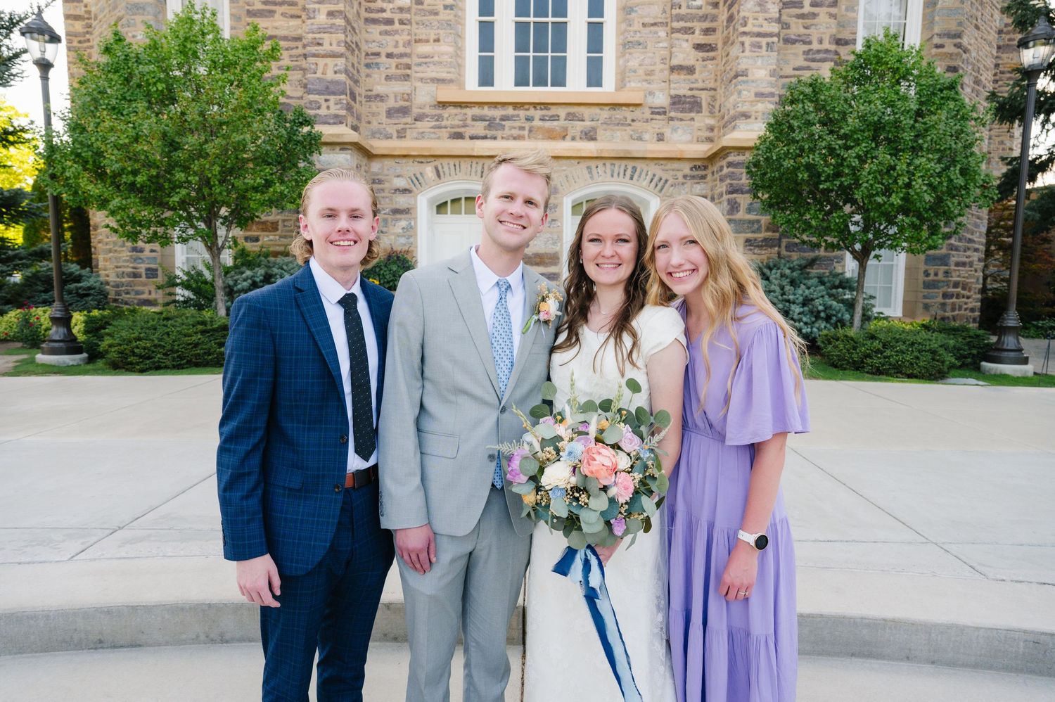 A group of four people pose for a formal photo outside a stone building on a wedding day.