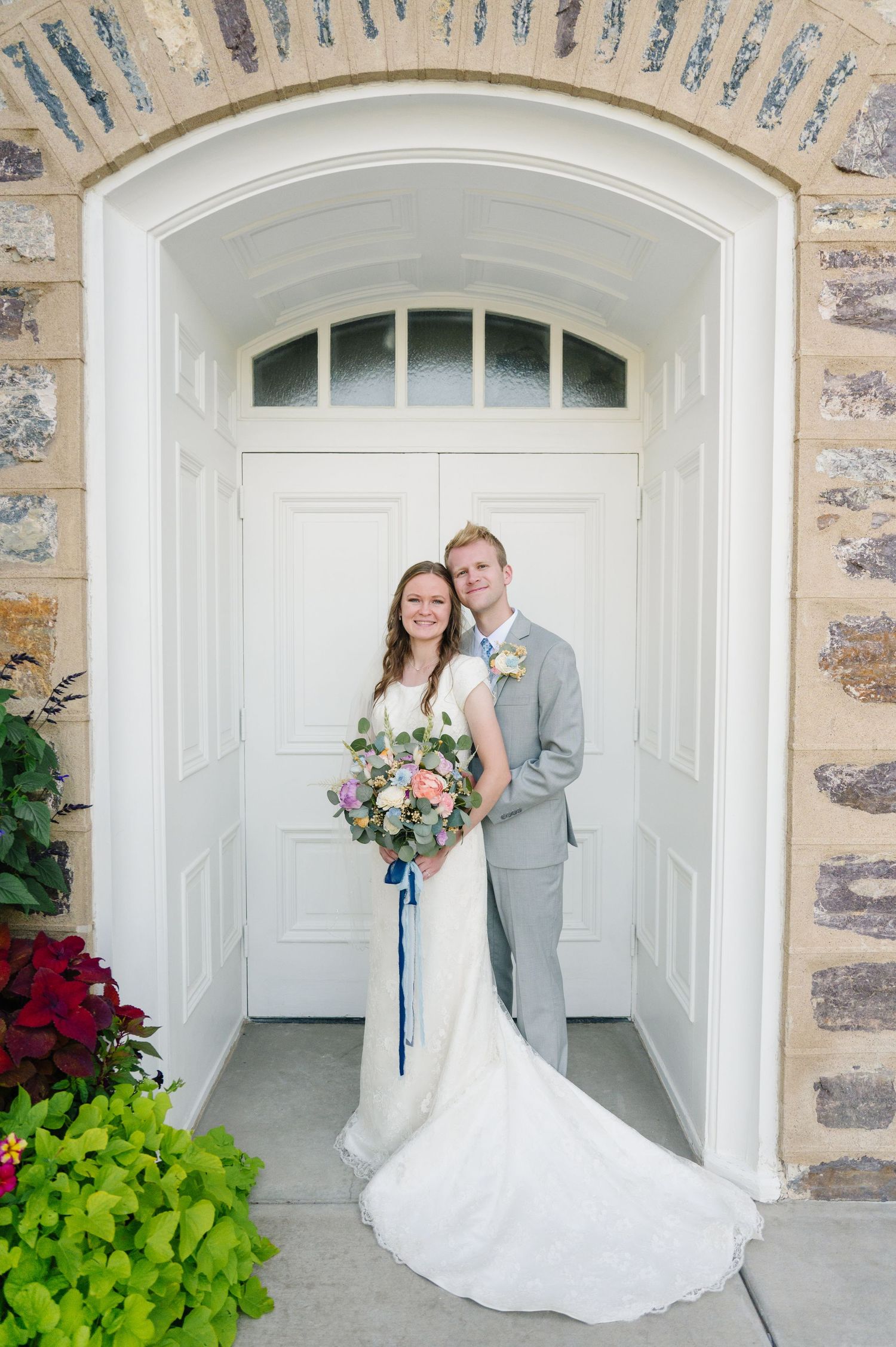 A couple poses in formal attire under a stone archway entrance with flowers.