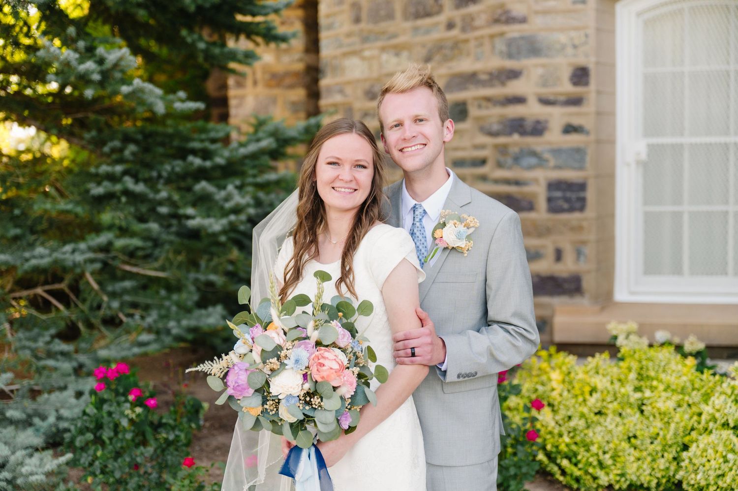 A couple in wedding attire poses in front of a stone building with blooming garden.
