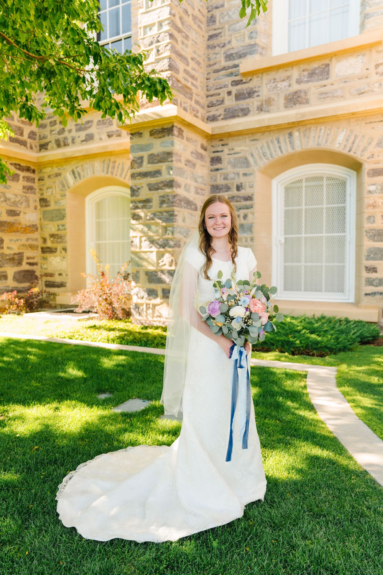 A bride in a classic white wedding gown with a trailing veil poses in front of a stone building with arched windows.
