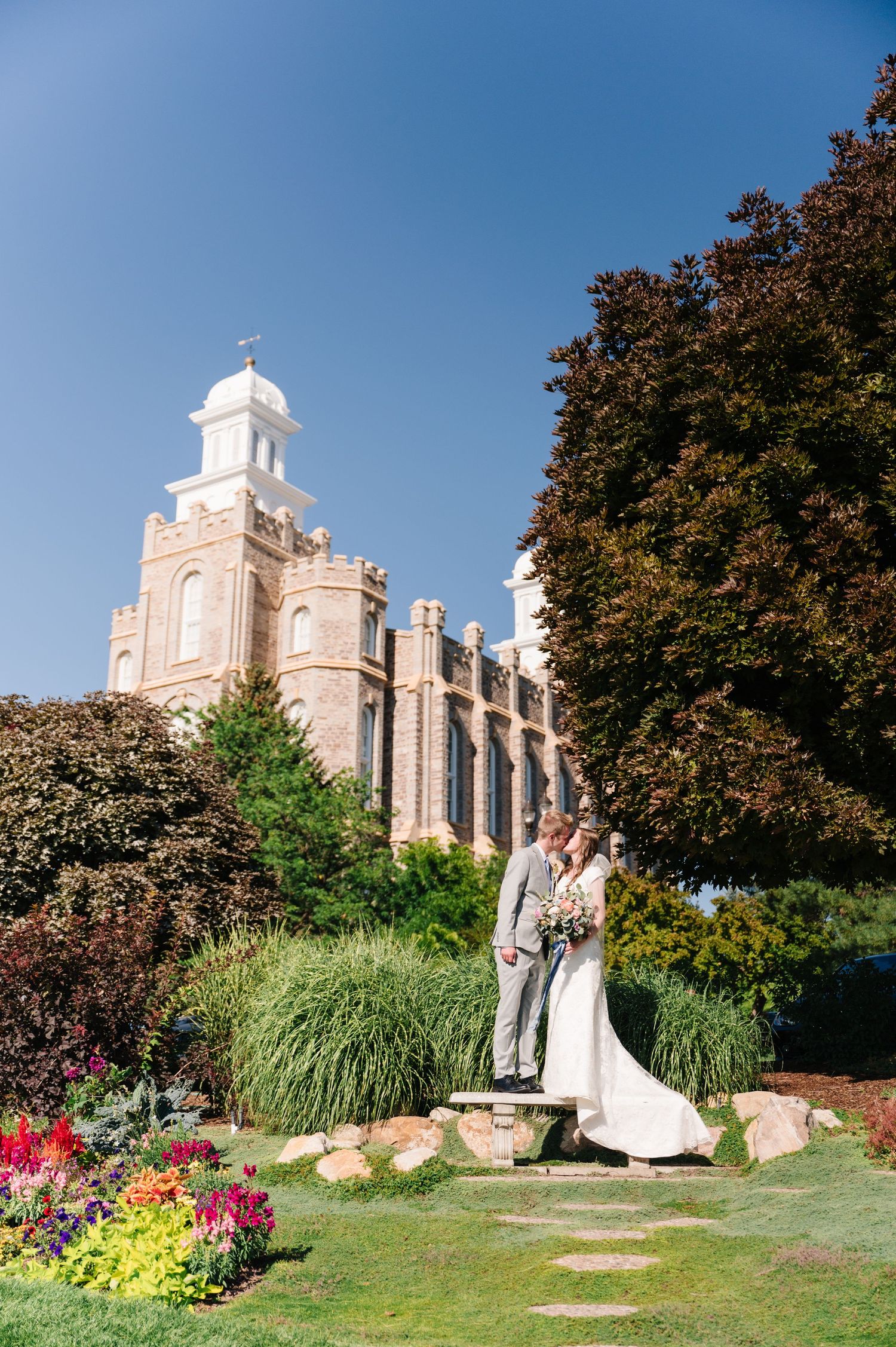 Wedding couple standing in a garden near a grand white stone temple building with colorful flowers in bloom.
