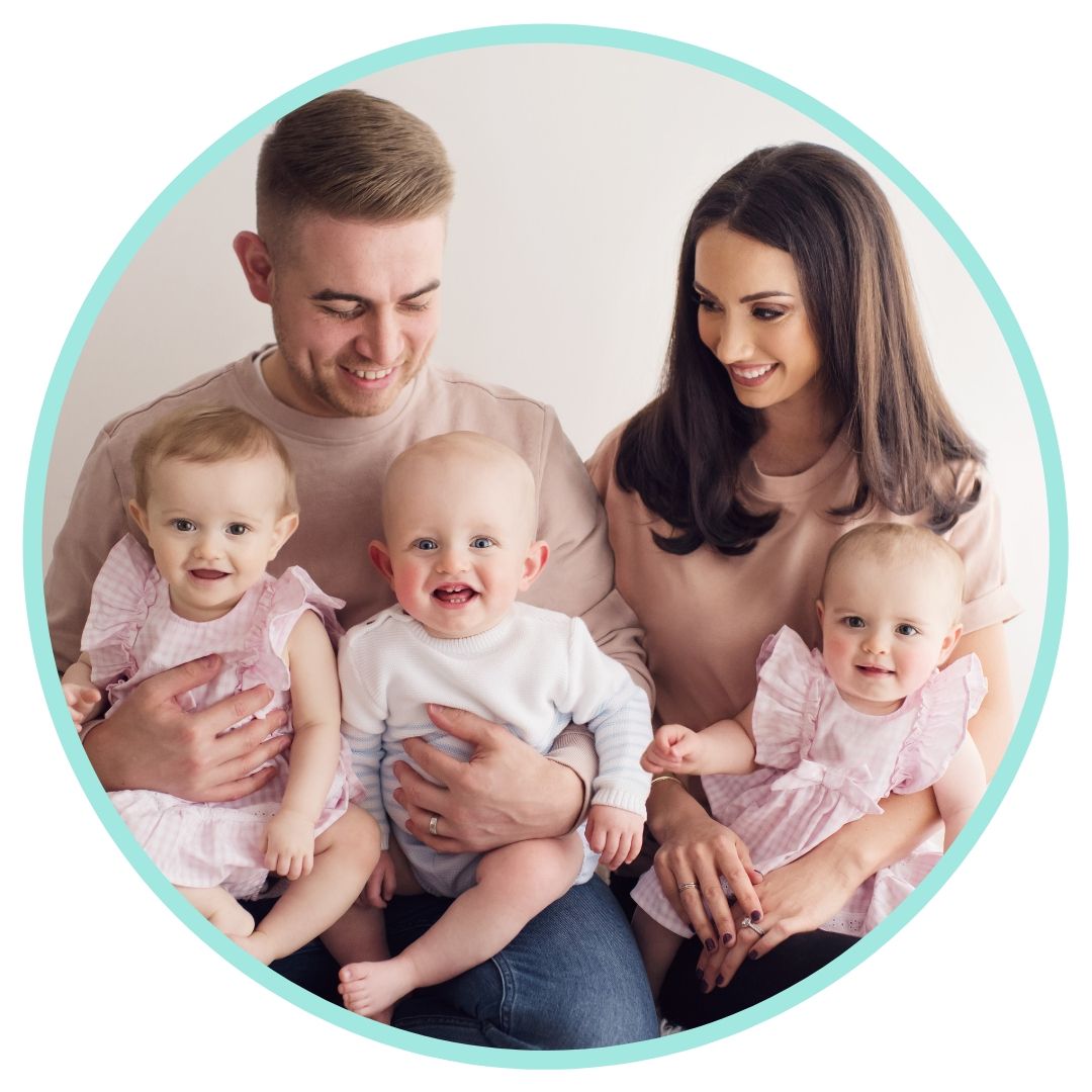 A family with three young babies in matching pink outfits sits together on a white background.