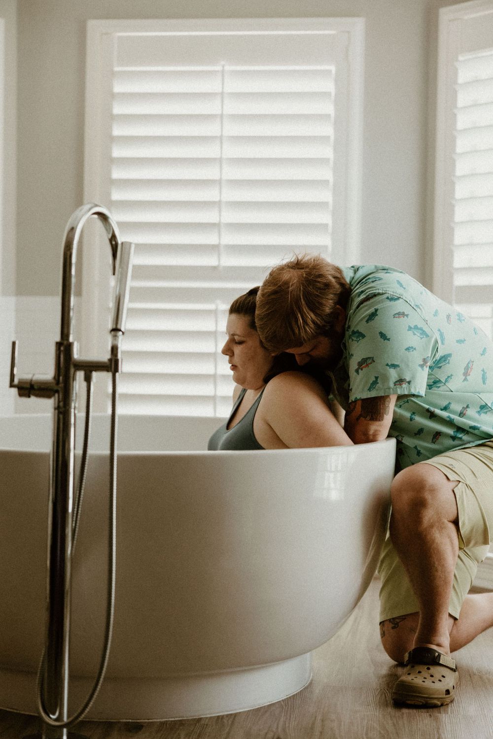 Two people sharing an intimate moment near a modern freestanding bathtub with window shutters.