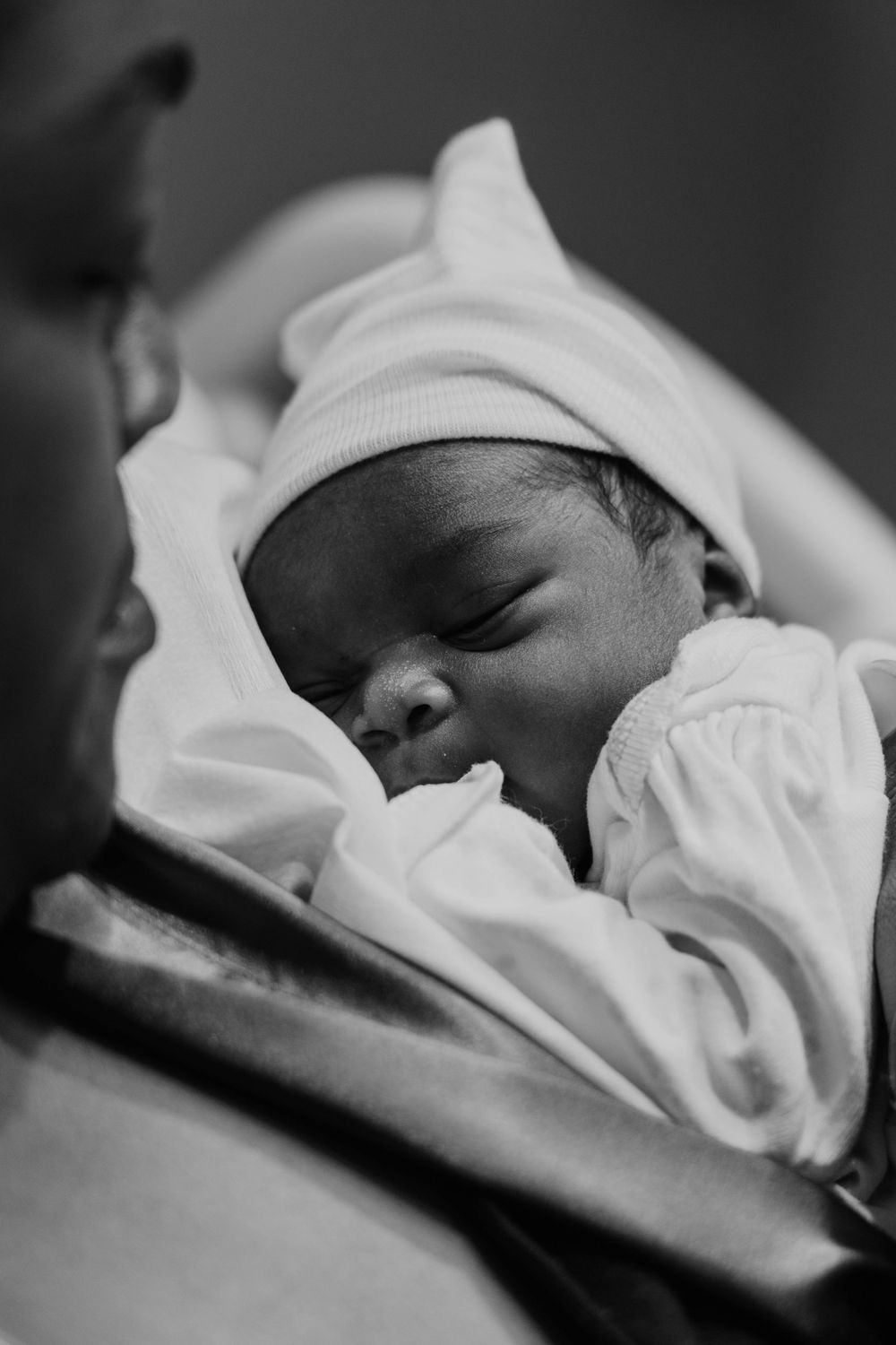 A newborn baby sleeps peacefully wrapped in white blankets with a matching white cap.