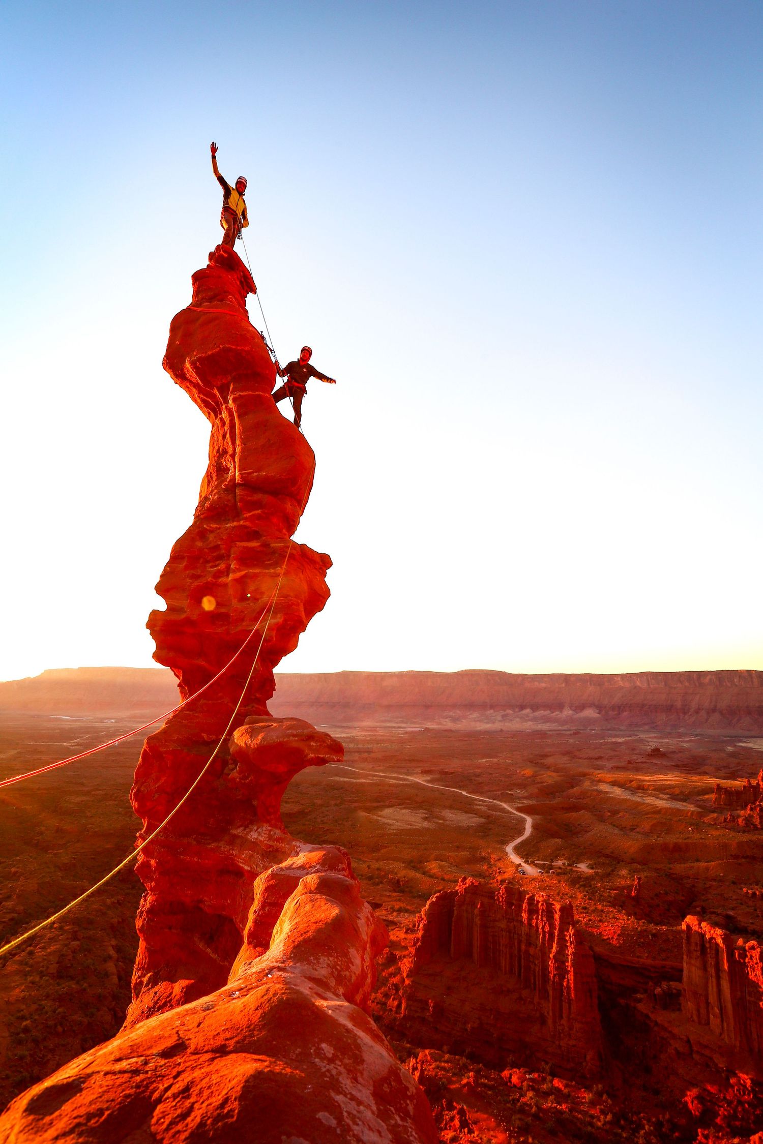 Two rock climbers scaling a tall red sandstone spire at sunset in a vast canyon landscape in Utah.