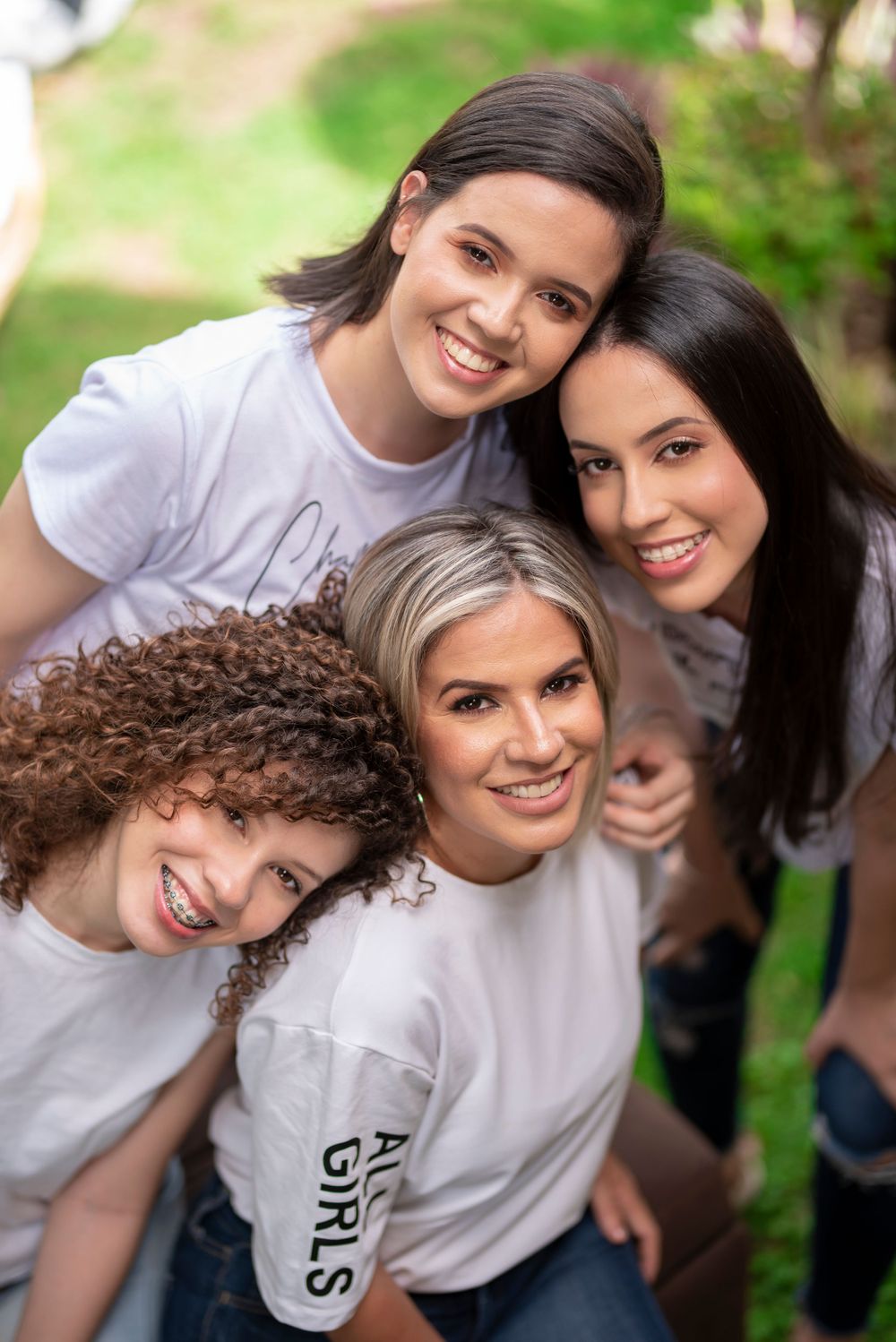 Four smiling women in matching white shirts posing together outdoors in a joyful group portrait photo.