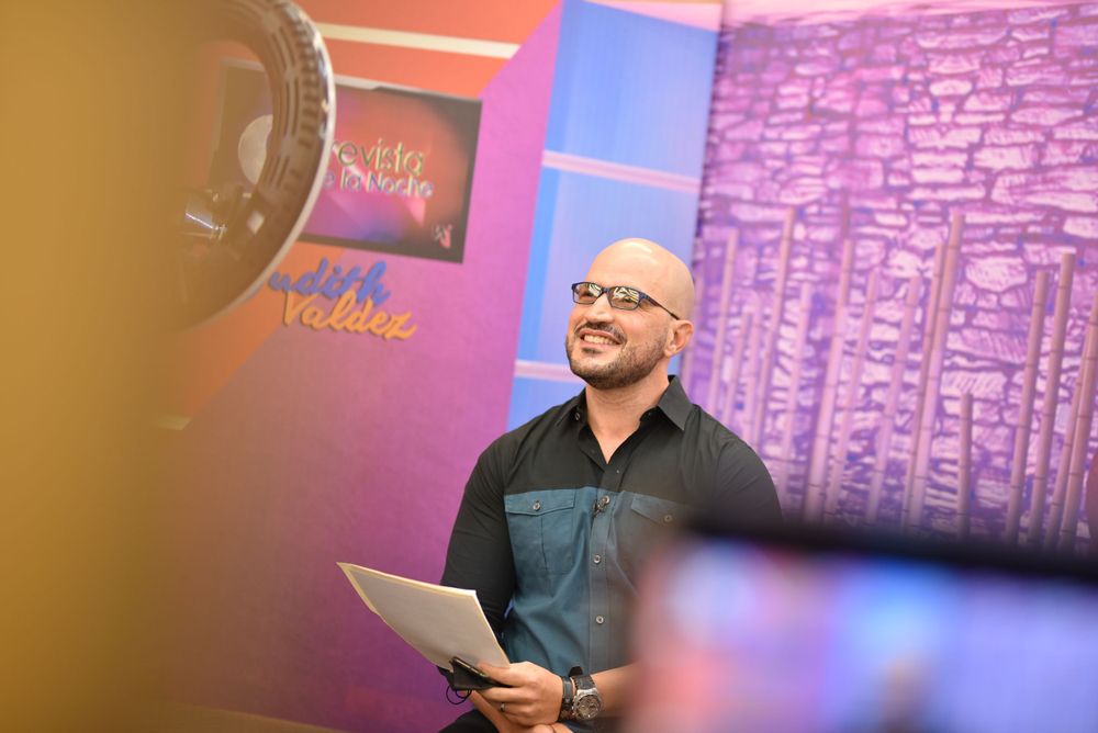 Smiling bald man holding papers standing in colorful TV studio broadcast set background.
