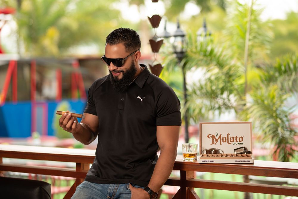 Stylish man in black polo wearing sunglasses checking phone at outdoor bar with whiskey.