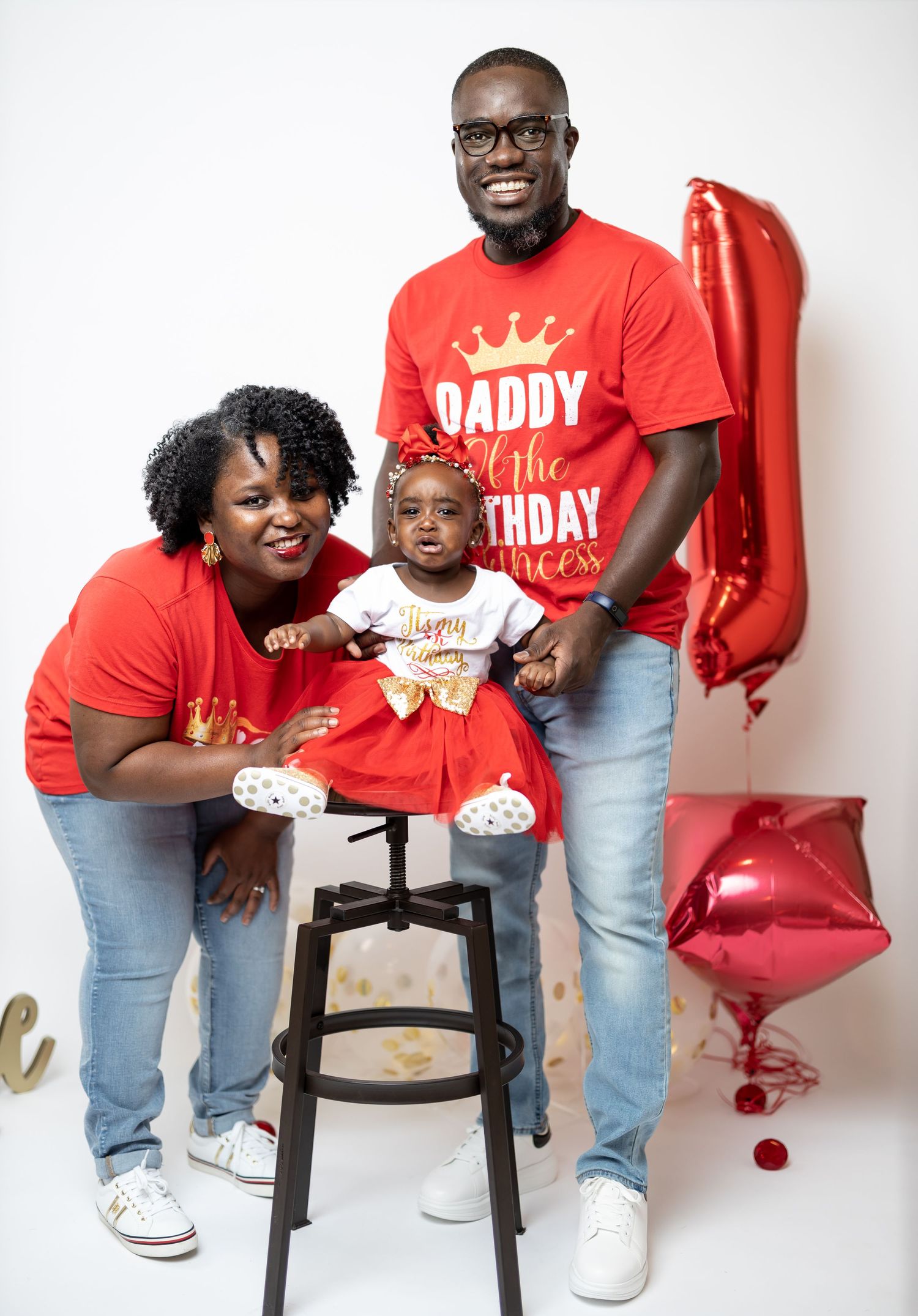 Family poses together by red balloons with child seated on wooden stool against white backdrop.