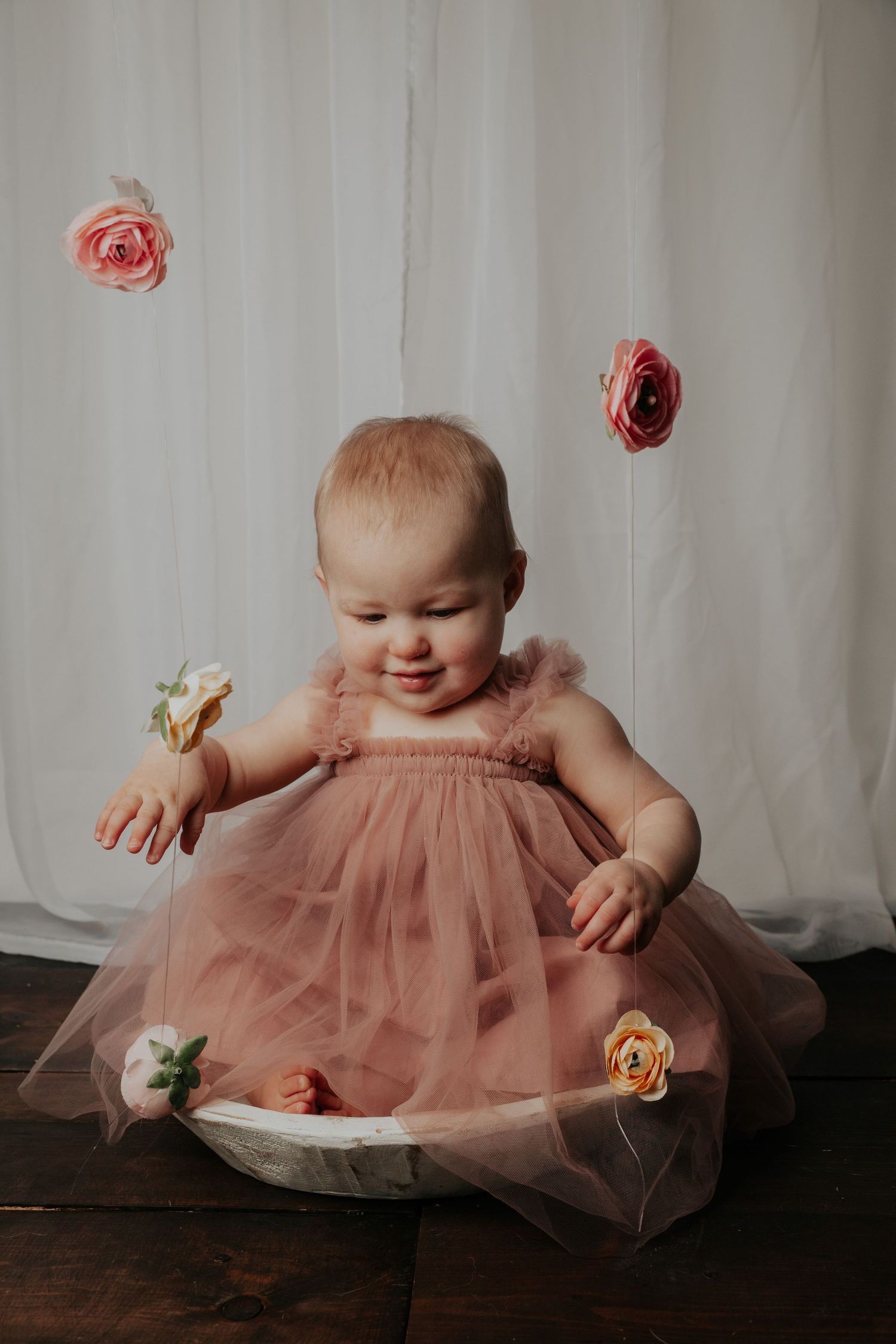 Baby wearing pink tulle dress plays with flowers on dark wooden floor.