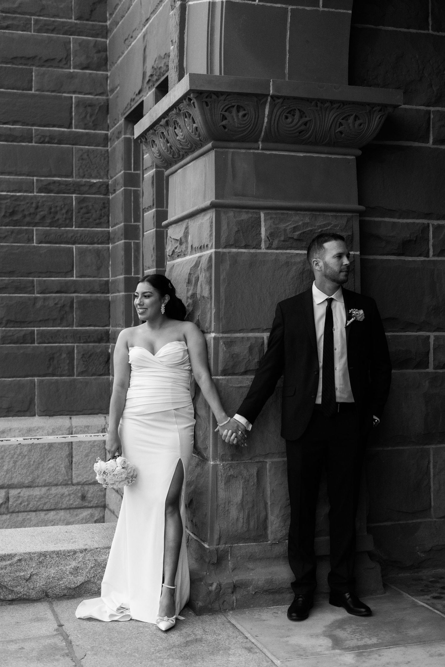 Stylish bride and groom hold hands while posing against historic brick building.