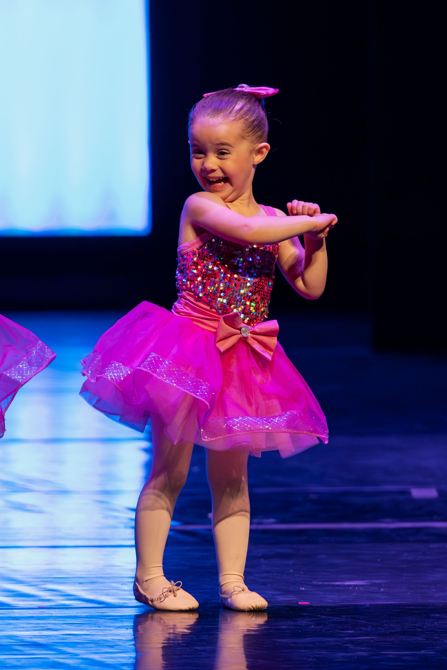 Small dancer in pink sparkly tutu performs on stage under dramatic lighting.