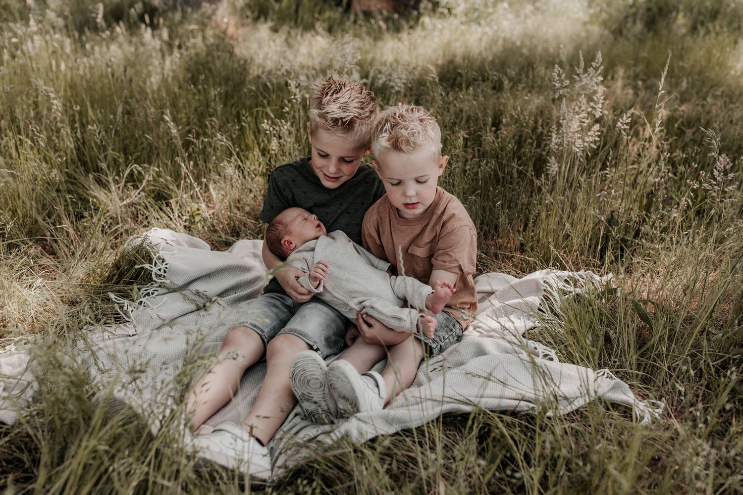 Lifestyle shoot buiten, broertjes zitten op een kleedje met hun nieuwe baby broertje op hun schoot