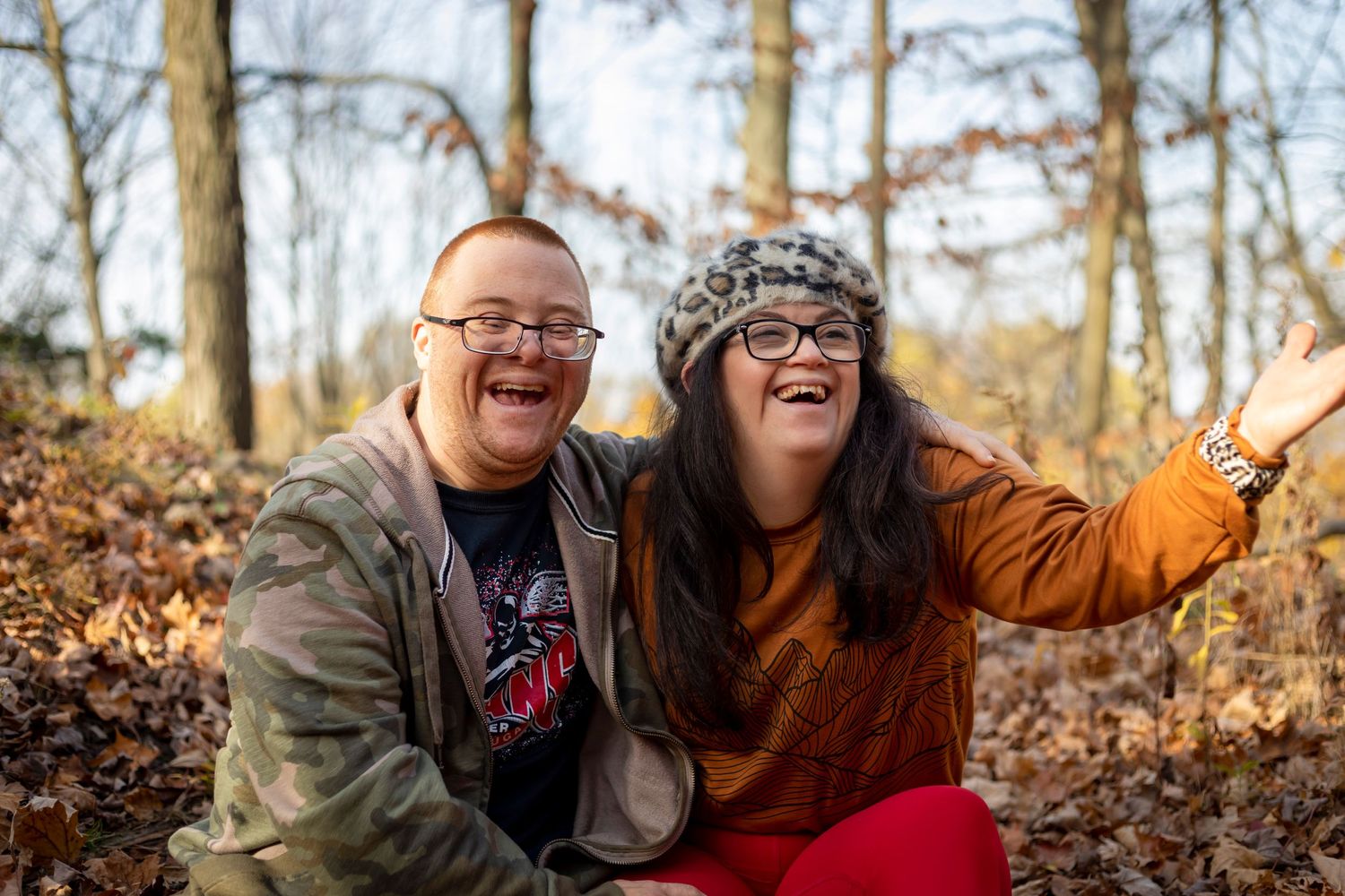 Two people laugh joyfully while sitting among fallen autumn leaves in the woods.