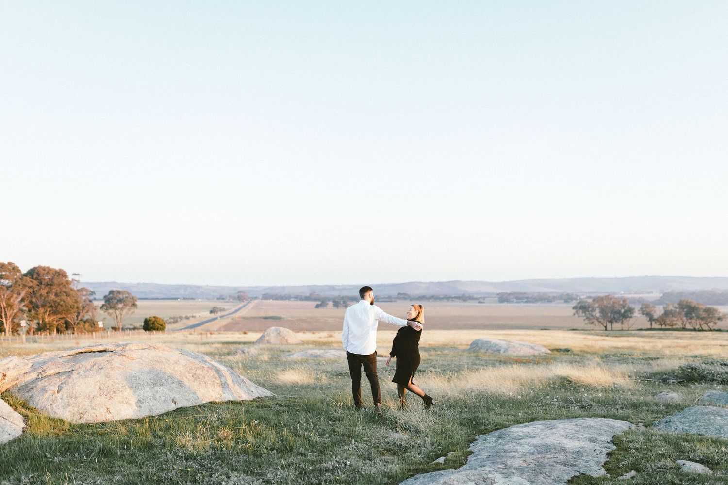 A couple strolls along a hilltop trail overlooking a vast pastoral valley.