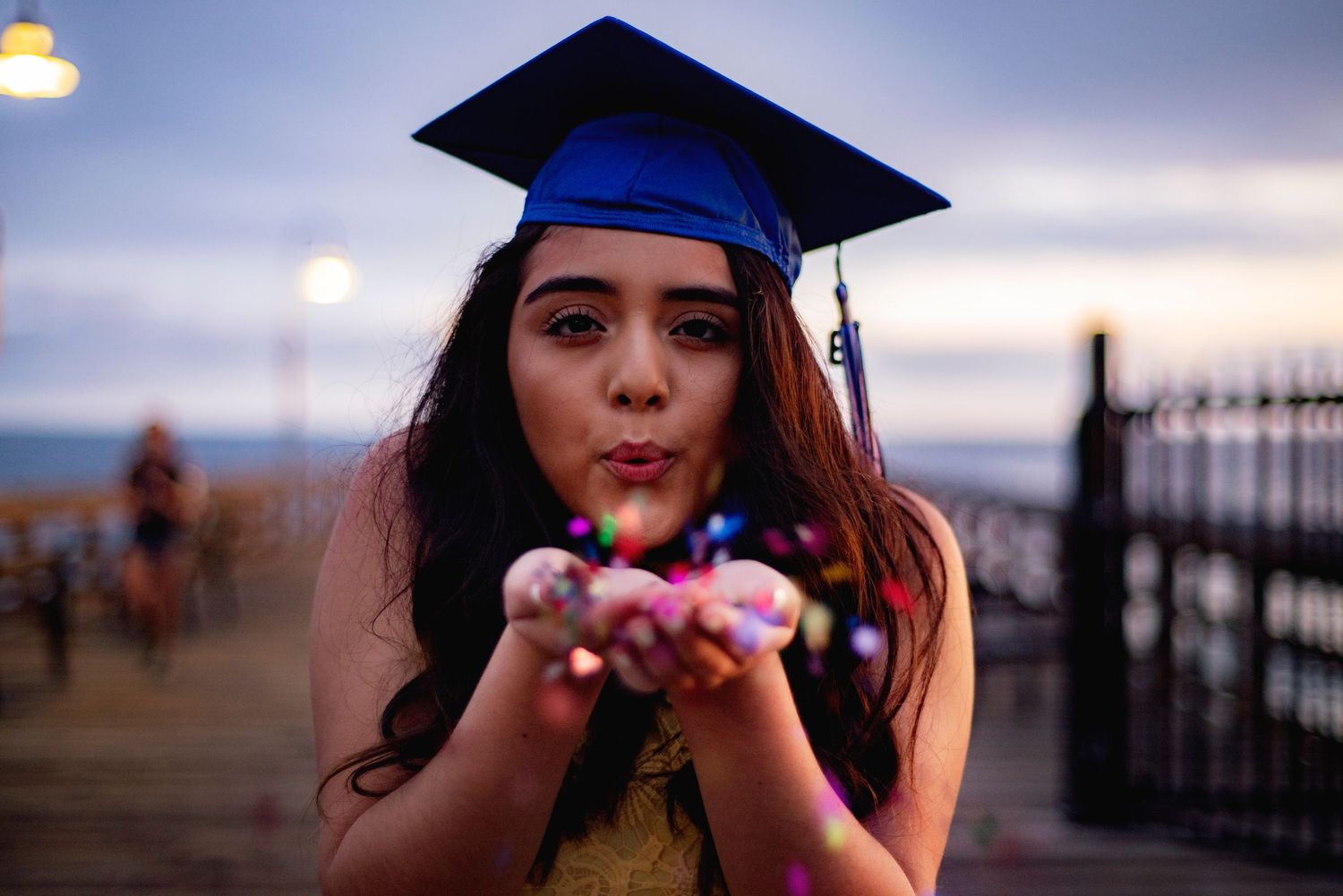 Graduate in blue cap and gown blowing glitter from hands at sunset.