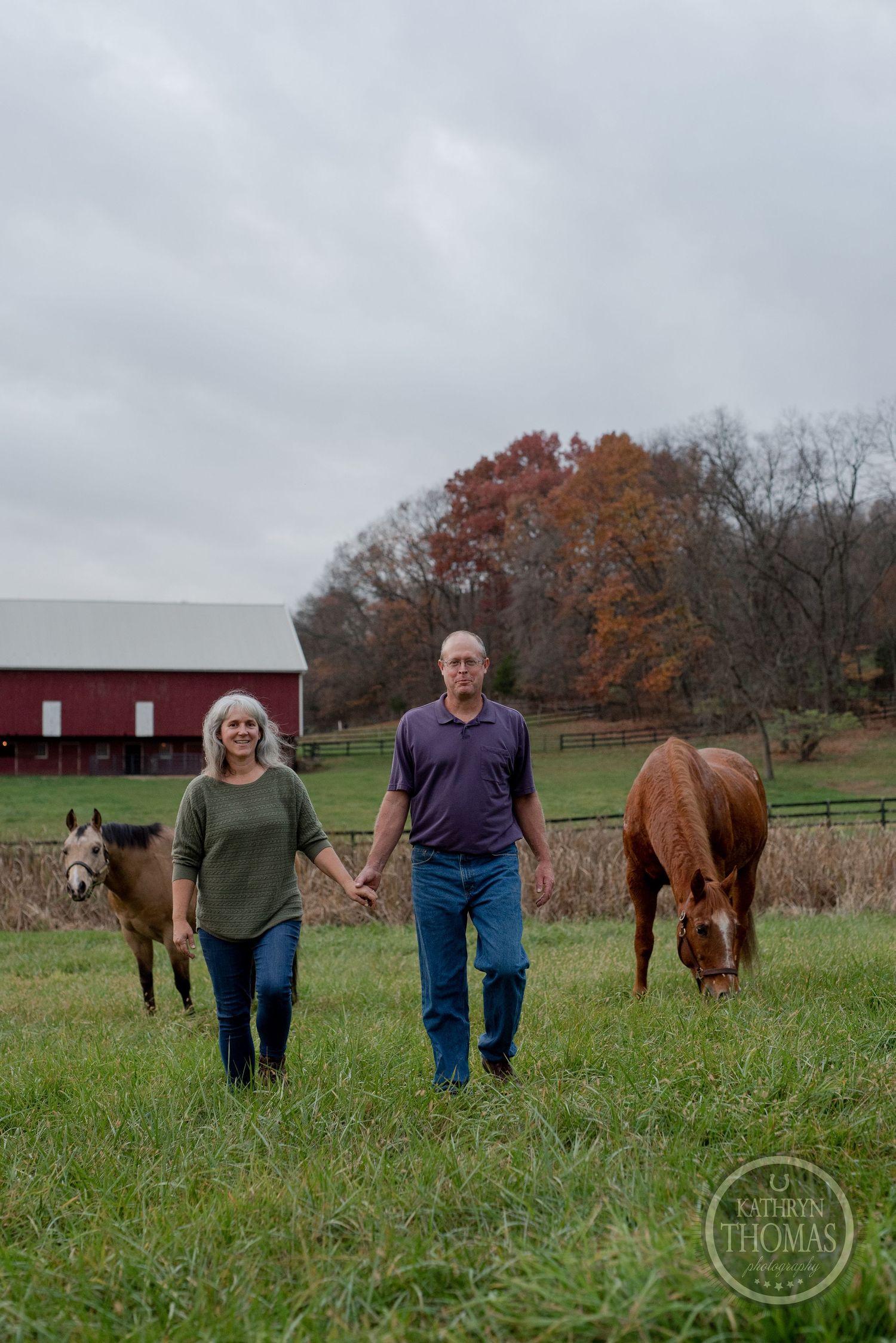 A couple walks with their horses in a grassy field with a red barn and autumn trees in the background.