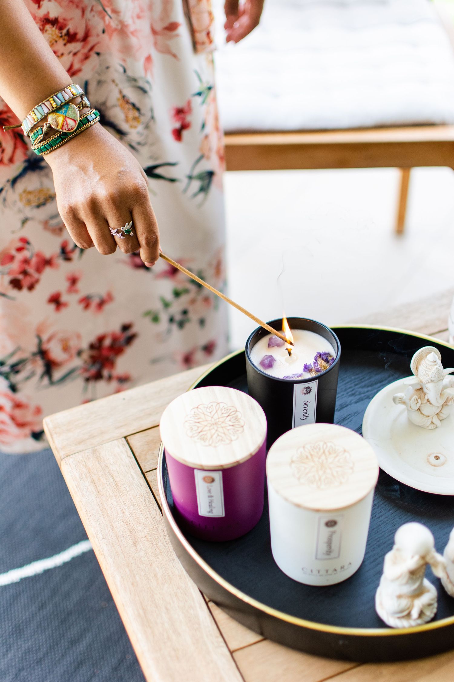 Person lighting decorative candles on wooden tray wearing floral dress and stacked bracelets.