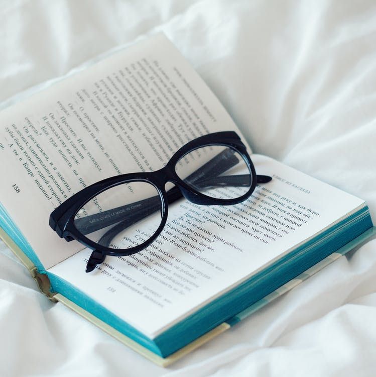 Black reading glasses resting on open turquoise book against white bedding.