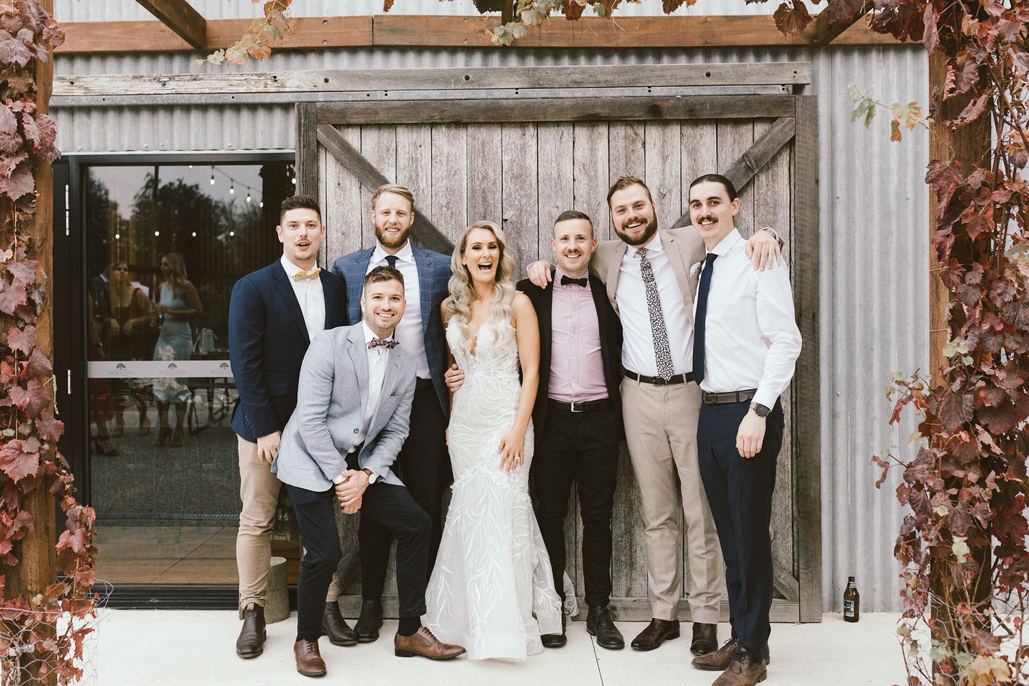 Wedding party posing together in front of rustic metal door framed by autumn foliage.