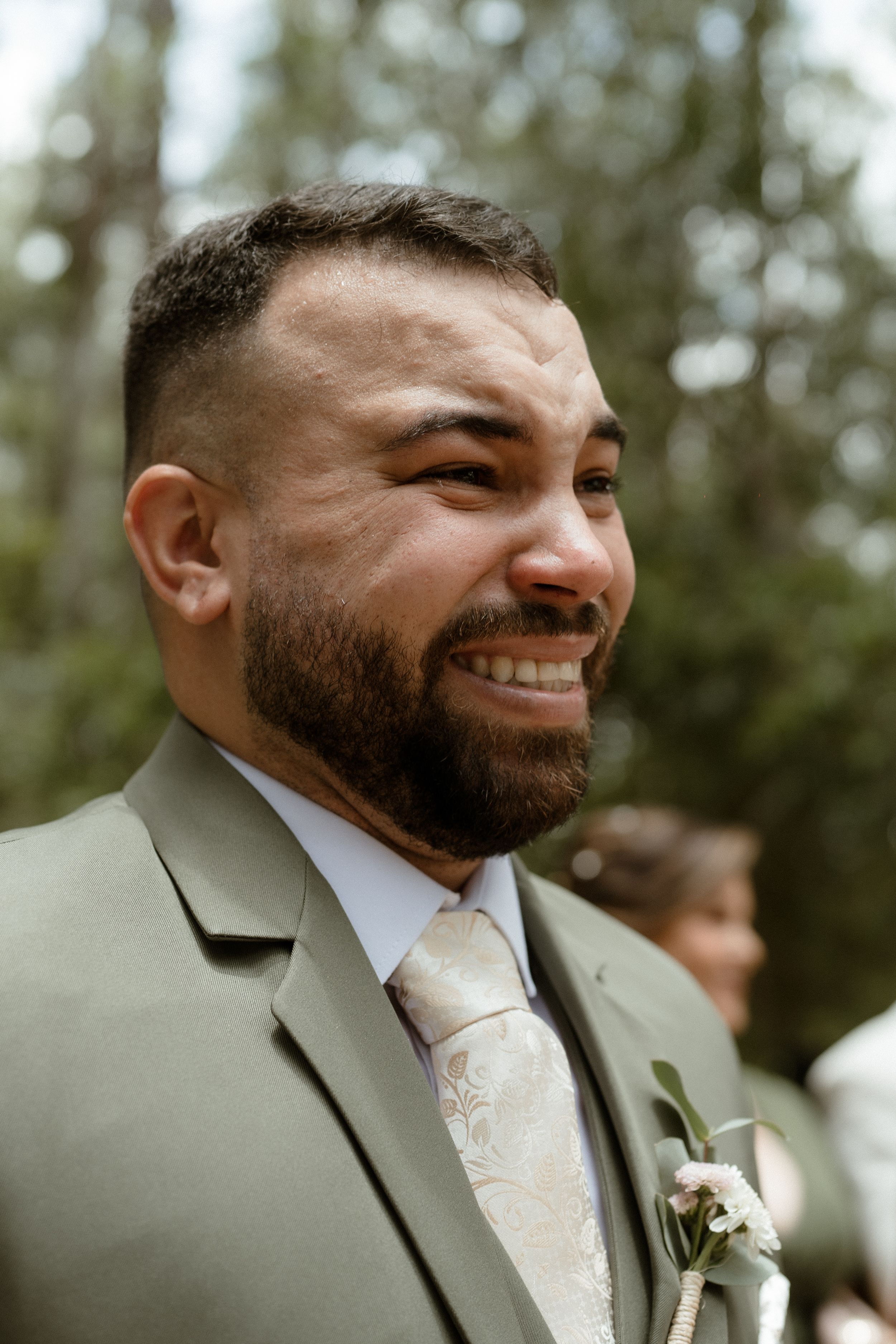 Groom in light gray suit with matching tie smiles during outdoor forest ceremony.