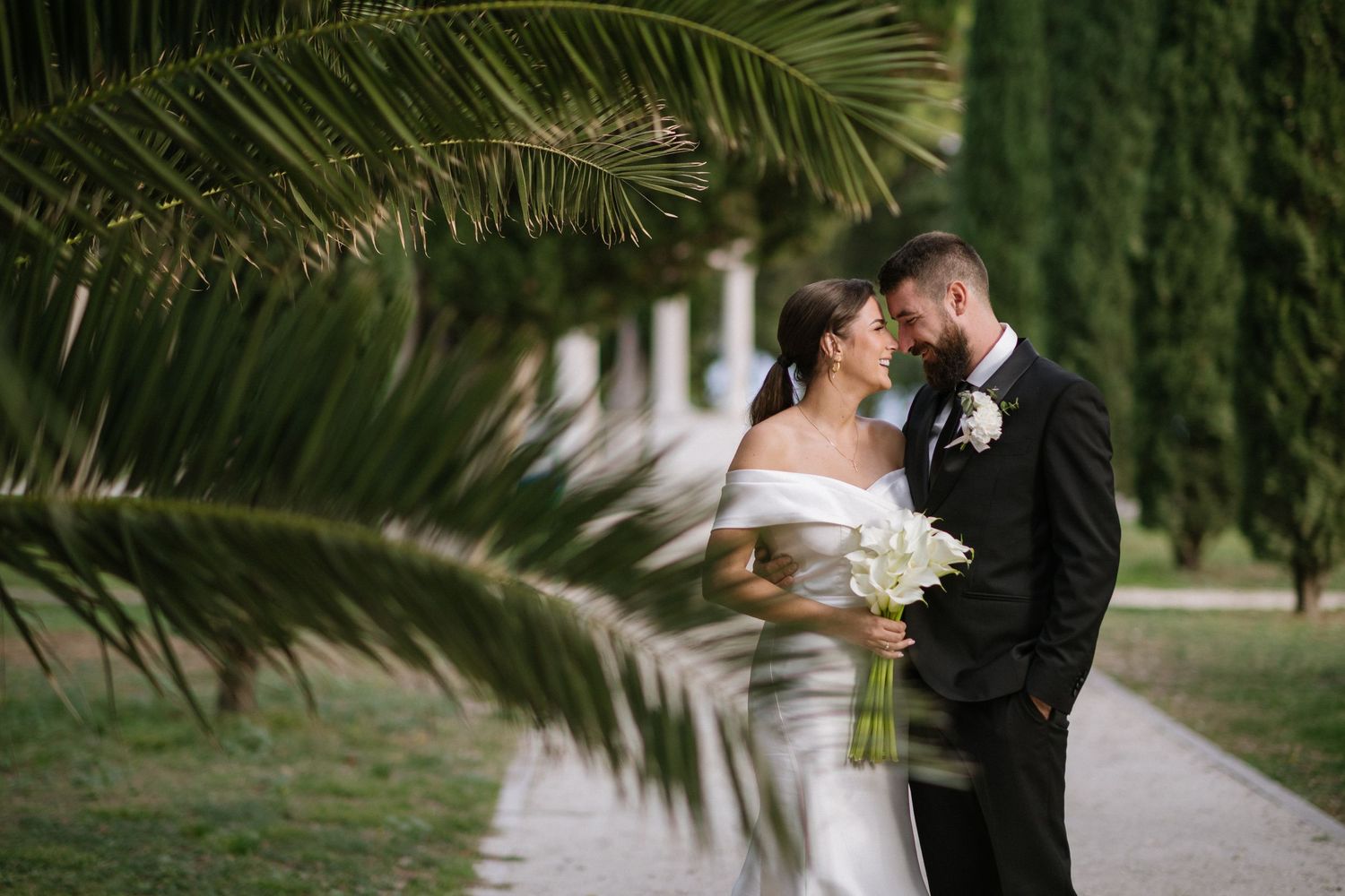 A couple shares a romantic moment under a palm frond along a garden path while holding a white bouquet.