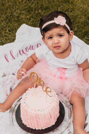 A baby in a pink tutu sits next to a decorated first birthday cake.