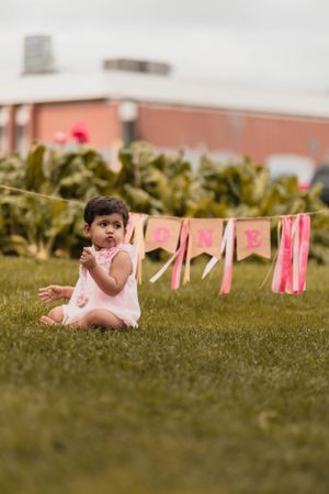 Young child sitting on grass with pink party decorations hanging on clothesline in background.